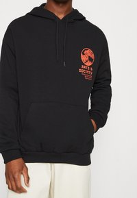 Sudadera negra con un bolsillo canguro frontal, capucha con cordones y texto gráfico naranja en el pecho. Material de algodón, ajuste estándar.