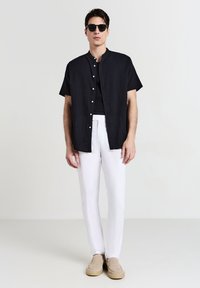 Schwarz kurzärmliges Hemd mit Knopfleiste, weiße Hose, beige Slipper und schwarze Sonnenbrille. Glattes Material mit lässiger Passform.