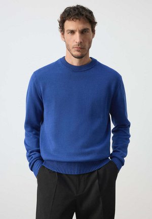 Hombre con cabello corto y rizado que lleva un suéter azul de cuello redondo y pantalones negros, de pie con las manos en los bolsillos frente a un fondo liso.