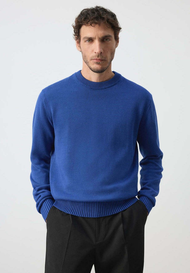 Homme aux cheveux courts et bouclés portant un pull bleu à col rond et un pantalon noir, debout les mains dans les poches devant un fond uni.