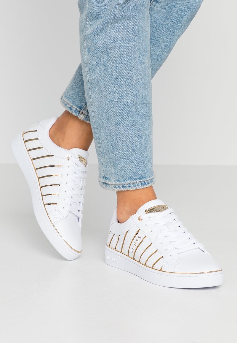 Baskets basses blanches avec des rayures verticales dorées portées avec un jean denim bleu clair sur un fond neutre.