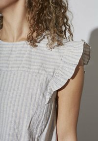 Lyseblå og hvid stribet top med flæseærmer. Fremstillet af struktureret stof, har en løs pasform og bølgende kant.