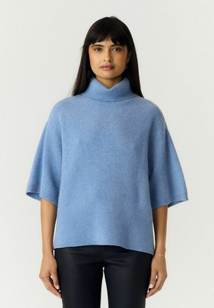 Une femme porte un pull bleu clair oversize à manches courtes et à col roulé, ainsi qu’un pantalon noir, sur un fond uni.