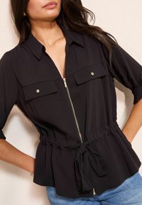 Melnā aizdare blūze ar apkakles izgriezumu, divām krūšu kabatām un savāktu jostasvietu ar sasienamu elementu. Mīksts audums, gluda tekstūra.