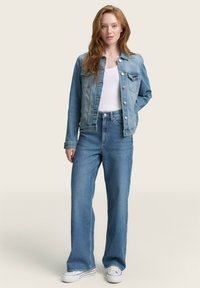 Svetlo modra denim jakna s sprednjimi gumbi, žepoma na prsih in dolgimi rokavi, v kombinaciji z visokim pasom svetlo modrimi kavbojkami in belimi športnimi copati.