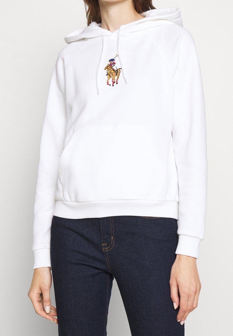 Personne portant un sweat-shirt à capuche blanc avec une poche kangourou avant et un petit logo brodé de joueur de polo, assorti à un jean bleu foncé.