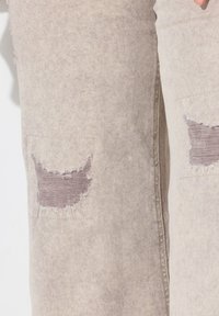 Jeans in denim beige con toppe sfilacciate all'altezza delle ginocchia e una finitura leggermente texturizzata. Cuciture visibili lungo le cuciture e un aspetto sbiadito.