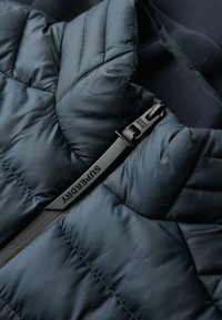 Granatowa puchowa kurtka o pikowanej strukturze, z czarnym suwakiem z napisem "Superdry" oraz miękką podszewką z materiału.