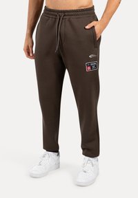 Hnědé tepláky s elastickým pasem, bočními kapsami a detailem s logem. V kombinaci s bílými teniskami. Hladká textilie.