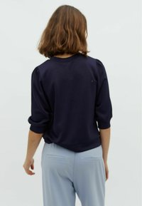 Pullover blu navy con scollatura rotonda e maniche a sbuffo, abbinato a pantaloni azzurri chiari con tasche posteriori. Texture liscia su tutta la superficie.