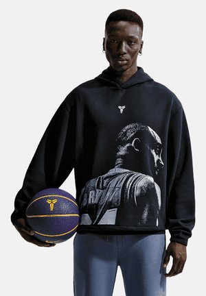 Hombre con sudadera negra con capucha con estampado en blanco y negro de jugador de baloncesto sostiene un balón de baloncesto en una mano, de pie frente a un fondo liso.