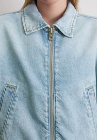 Eine Nahaufnahme einer Person, die eine hellblaue Jeansjacke trägt, mit einem Reißverschluss vorne und einem Kragen, vor einem neutralen Hintergrund.