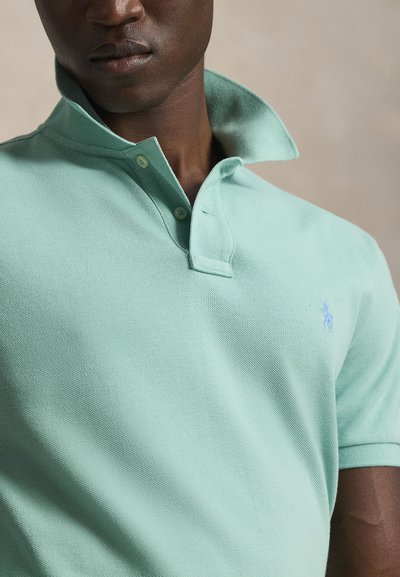 Κοντινό πλάνο ενός άνδρα που φοράει γαλαζοπράσινο πόλο πουκάμισο με σηκωμένο γιακά και μικρό κεντημένο λογότυπο στο στήθος.