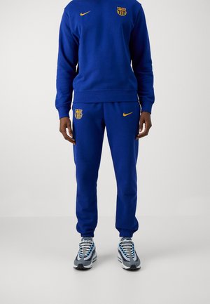 Persoon draagt een blauw Nike Barcelona trainingspak met gele logo's, gecombineerd met grijze en witte sportieve sneakers, staand tegen een effen achtergrond.