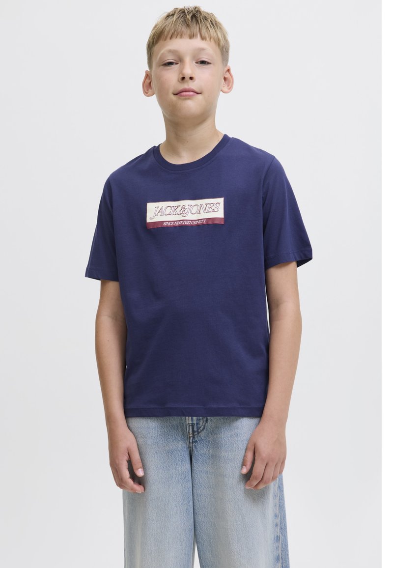 Garçon portant un t-shirt bleu marine Jack & Jones et un jean bleu clair, debout devant un fond blanc uni.