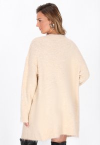 Beige oversize svetr s měkkou, huňatou texturou. Má dlouhé rukávy, žebrované manžety a decentní dekorativní vzor na rukávu.