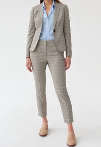 Ensemble composé d'un blazer et d'un pantalon à carreaux dans des tons beiges, offrant une coupe ajustée, une fermeture à un bouton, associé à un blouse bleu clair et des mocassins beige.