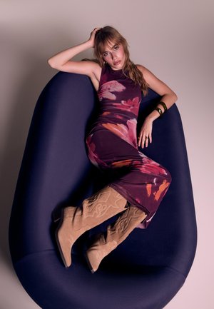 Frau, die sich auf einem tiefblauen modernen Stuhl zurücklehnt, trägt ein lila Blumenkleid und beige bestickte knielange Stiefel, mit der Hand am Kopf.