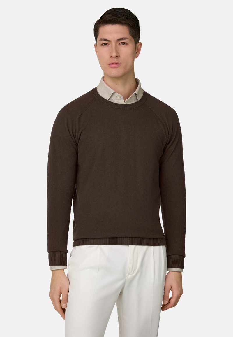 Homme portant un pull marron foncé à col rond sur une chemise beige à col, avec un pantalon blanc plissé, debout devant un fond uni.