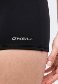 Κοντινό πλάνο του μηρού ενός ατόμου που φοράει μαύρο σορτς με το λογότυπο "O'NEILL" σε γκρι κοντά στο στρίφωμα, με το χέρι να ακουμπάει δίπλα στο πόδι.