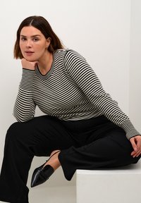 Gestreepte longsleeve top in zwart en crème, gecombineerd met wijde zwarte broek en puntige zwarte schoenen. Model zit op een witte voetstuk.