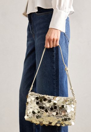 Mano sosteniendo un pequeño bolso de lentejuelas doradas con asa delgada, vistiendo jeans azules y camisa blanca de manga larga.