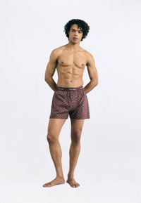 Sarkanas rakstainas bokseršorti ar melnu jostu, uz kuras redzams "Calvin Klein" logos. Audums izskatās mīksts un elpojošs. Modelis stāv basām kājām.