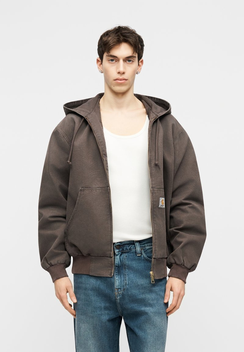 Giovane uomo che indossa una giacca con cappuccio marrone con cerniera sopra una canottiera bianca e jeans blu, in piedi contro uno sfondo chiaro uniforme.