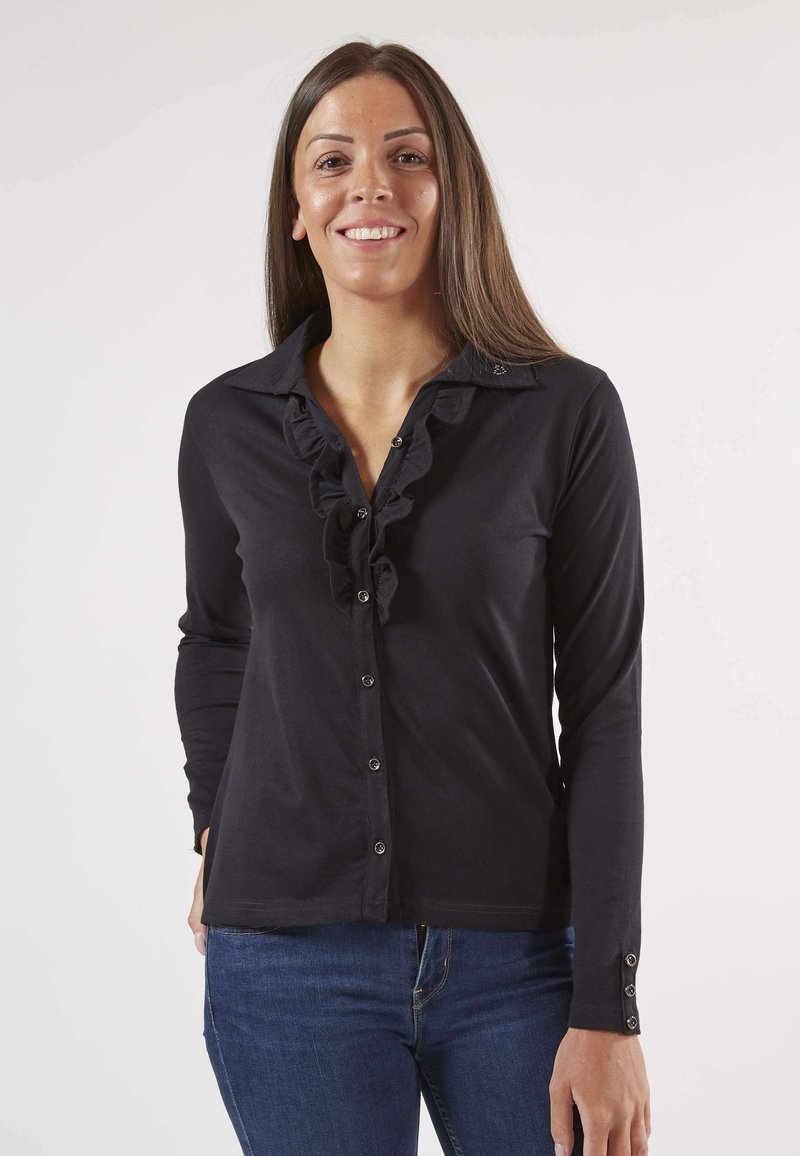 Chemise noire à manches longues avec un devant froncé, fermeture à boutons et coupe ajustée. Le tissu semble lisse avec une légère brillance. Portée avec un jean bleu.