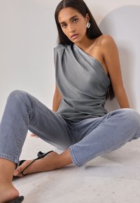 Graues One-Shoulder-Top mit drapiertem Stoff, kombiniert mit hellblauen Jeans. Schwarze Sandalen sichtbar, sitzend auf einer neutralen Oberfläche.