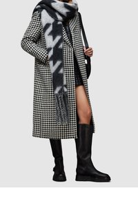 Schwarz-weißer Hahnentritt-Mantel mit tiefem Ausschnitt und seitlichem Schlitz, kombiniert mit einem grauen, gemusterten Schal und schwarzen kniehohen Stiefeln.