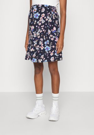 Kvinde iført en marineblå blomsterindpakket nederdel med hvide, pink og blå blomster, kombineret med hvide sneakers og hvide sokker med to grå striber.