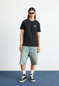 Schwarzes T-Shirt mit kurzen Ärmeln und einem weißen Logo auf der Brust, kombiniert mit hellblauen Denim-Shorts und schwarzen High-Top-Sneakers.