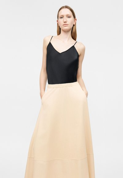 Γυναίκα με μακριά ίσια μαλλιά, ντυμένη με μαύρο τοπ χωρίς μανίκια και μπεζ φούστα με ψηλή μέση, με τα χέρια στις τσέπες, μπροστά σε απλό φόντο.