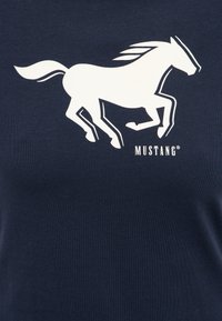 Silhouette d'un cheval mustang au galop en blanc sur un tissu bleu foncé avec le mot « MUSTANG » en dessous du cheval.