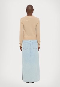 Personne aux cheveux courts portant un pull beige à manches longues et une jupe longue en denim bleu clair avec poches arrière et fente latérale, vue de dos.