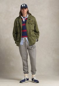 Grøn militærjakke, polo med røde striber, lyseblå skjorte, grå joggingbukser med logo, marineblå sneakers og NY-kasket. Afslappet lag-på-lag.