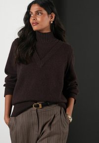 Dark brown knitted sweater with a high collar and V-neck design, paired with pinstriped trousers and a black belt with a gold buckle.