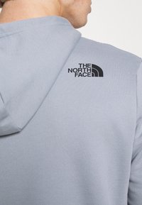 Sudadera con capucha de color gris claro y textura suave, con un logo negro de "The North Face" en la parte superior de la espalda, que contrasta con el color de la tela.