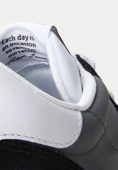 Nahaufnahme eines schwarz-weißen Sneakers mit Fokus auf den gepolsterten Kragen und das innere Stoffetikett mit gedrucktem inspirierendem Text.