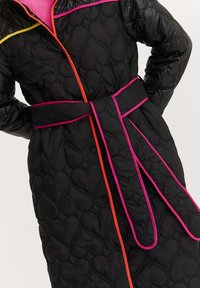 Čierny prešívaný kabát so vzorom srdca, zdobený ružovým, oranžovým a žltým lemovaním. Má opasok v páse a zips na zatváranie.