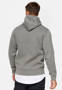 Grijze hoodie van gestructureerde breistoft, met ribgebreide manchetten en zoom, en een opvallende capuchon. Draagt men over een wit shirt.
