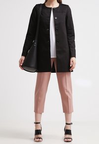 Nainen, joka on pukeutunut mustaan napitettuun takkiin, valkoiseen yläosaan, lyhyisiin vaaleanpunaisiin housuihin, mustiin korkokenkiin ja kantaa suurta mustaa olkalaukkua.