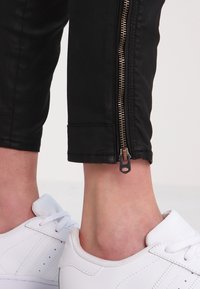Pantalon noir longueur cheville avec fermeture éclair sur le côté, porté au-dessus des chevilles, assorti à des baskets blanches à lacets.
