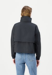 Kratka črna jakna z visokim ovratnikom, dolgimi rokavi in plastveno zasnovo na hrbtu. Material se zdi gladek z mat teksturo.