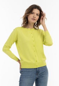 Cardigan a maglia di un brillante giallo, con chiusura a bottoni, polsini e orlo a costine e colletto rotondo. Indossato con jeans blu.