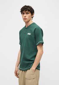 Νεαρός άντρας με κοντά σγουρά μαλλιά φοράει πράσινο πουκάμισο με κοντά μανίκια και μπεζ παντελόνι καμπαρντίνας, στέκεται μπροστά σε απλό φωτεινό φόντο.