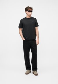 Νέος άνδρας με μαύρο t-shirt, μαύρο παντελόνι με φαρδιά φάσα, μπεζ μπότες και μαύρα γυαλιά ηλίου, στέκεται μπροστά σε λευκό φόντο.