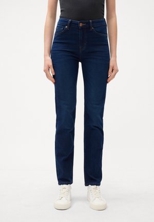 Personne portant un jean bleu foncé taille haute à jambes droites et des baskets blanches, debout devant un fond clair uni.