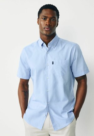 Man wearing light blue short-sleeve button-up shirt with chest pocket and beige pants, standing with hands in pockets against white background.